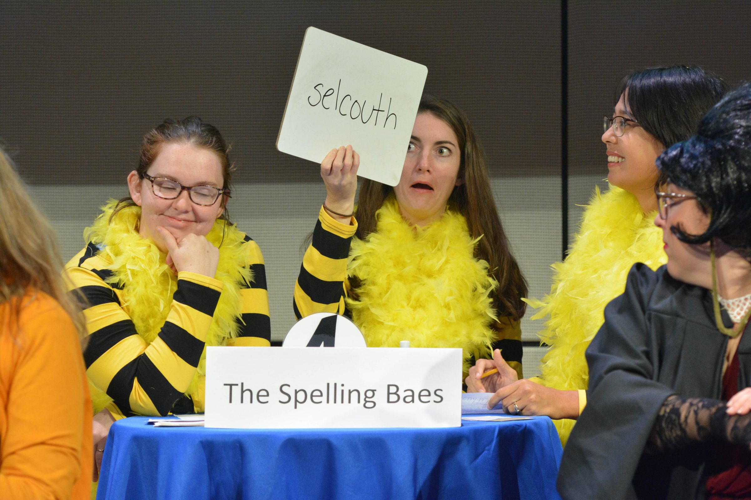 2018 Spelling Bee contestants "The Spelling Baes" stand at a table on the Yale School of Management stage, laughing and smiling as one of their teammates hold up a small white board with the word "selcouth."
